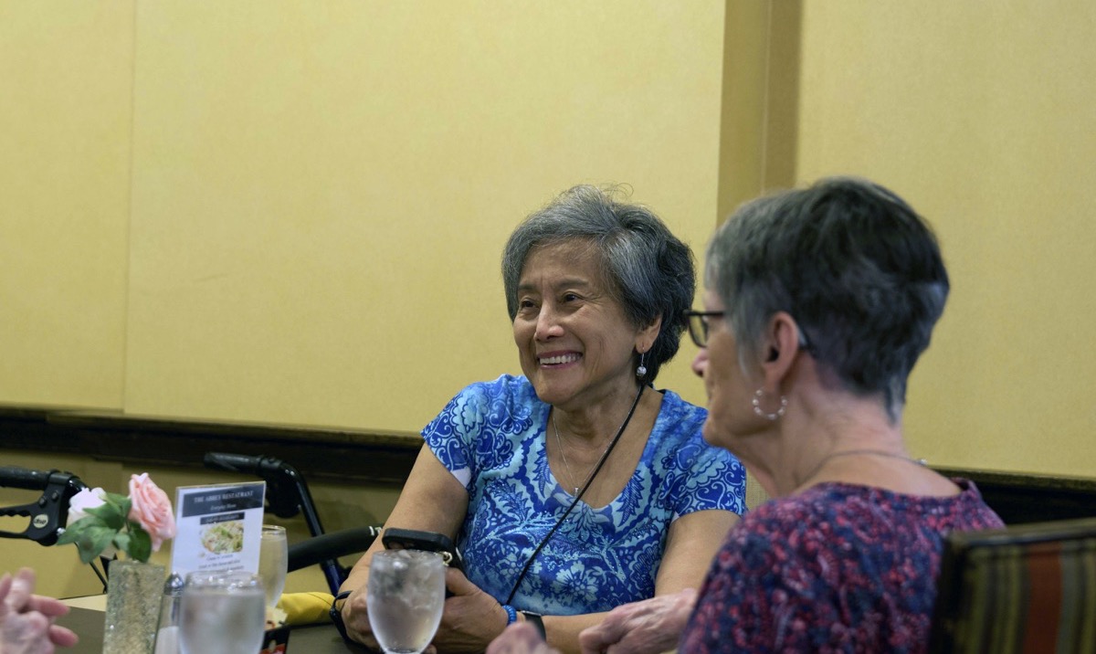 Two senior residents smiling and talking together over lunch in the dining room at The Abbey at Westminster Plaza